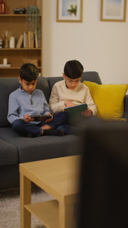 Vertikales-Video-Von-Zwei-Kleinen-Jungen,-Die-Zu-Hause-Auf-Dem-Sofa-Sitzen-Und-Spiele-Spielen-Oder-Auf-Ein-Digitales-Tablet-Oder-Ein-Tragbares-Spielgerät-Streamen